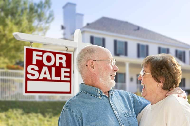A senior couple standing in front of their house with a for sale sign in the background.